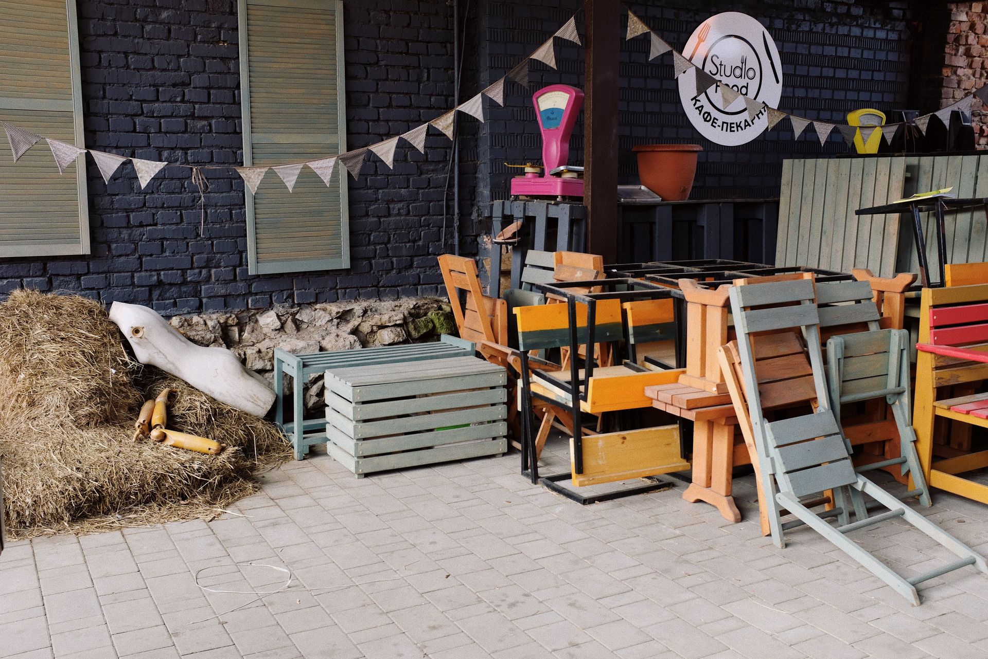 Street paved yard of small cafe with folded wooden chairs and benches placed on each other and body of mannequin on haystack near brick wall